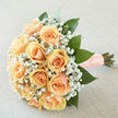 Soft peach roses arranged in a perfect, rounded cluster form the heart of this bouquet, their creamy-apricot petals curling in classic, velvety spirals that seem lit from within under gentle daylight. Nestled around and between them, clouds of white gypsophila create an airy, enchanted frame, their tiny blossoms scattering soft highlights across the composition. Large, deep emerald leaves provide contrast and structure, placed with an experienced hand so the bouquet reads as both lush and composed. The stems are tightly wrapped in a textured peach ribbon trimmed with pearls, a tactile detail that lends vintage romance and a finished, personal quality to the piece. The overall light is diffuse and flattering, revealing subtle creamy undertones in the petals and the nuanced green of the foliage; the scent is equally restrained-fresh green notes and the delicate sweet breath of roses with a faint lift from the gyp. As a Mill Hill florist I often prepare similar hand-tied bouquets for couples marrying near Mill Hill Broadway station or for small ceremonies in the Mill Hill Village greens, where that quiet, refined palette feels especially at home. Available from Flowers Mill Hill for local delivery, this arrangement carries an emotional tone of anticipation and devoted warmth, suited to bridal parties, anniversary moments, or intimate celebrations. It's crafted to be held close-comforting, elegant, and enduring-so each person who receives it remembers the gentle care that went into every layered petal and ribboned stem.