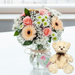 On a brilliant white tabletop, a tender anniversary gift ensemble is carefully positioned, the clarity of the setting allowing every detail to stand out as if in a Mill Hill studio flooded with daylight. Central to the scene is a rounded bouquet in a clear glass jar vase, the transparency revealing straight green stems gathered neatly in the water. The flowers form a gentle pastel palette: soft pink roses open in layered curls, peach gerbera daisies fan out around rich dark brown centres, and neat white chrysanthemums with hints of green at their cores add crisp brightness. Wispy stems of white baby's breath are tucked throughout, lending a light, airy veil around the more prominent blooms. A length of natural, slightly rough twine encircles the neck of the jar and is tied into a simple bow, from which a small pale gift tag dangles. The tag is decorated with a pink floral or heart illustration and subtle green handwriting, giving the impression of a lovingly handmade, personalised gesture from a local Mill Hill florist. To the right of the vase, a small cream teddy bear sits upright, turned gently toward the flowers. Its soft, textured fur, dark eyes, brown button-like nose, and slim brown satin ribbon at the neck make it look comforting and nostalgic, like a keepsake waiting to be cuddled. The background is a wash of bright, blurred whites and soft tones, suggesting daylight filtering through windows that might look out toward Mill Hill Park or nearby residential streets. The overall atmosphere is one of affection, reassurance, and celebration, ideal for marking anniversaries, birthdays, or simply saying I'm thinking of you within the Mill Hill community.