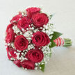 A luxurious bouquet of deep red roses and white baby's breath is showcased in serene, natural light, suggesting a peaceful Mill Hill afternoon where time seems to slow. The roses are the focal point, each bloom richly saturated in crimson, with velvety petals that layer elegantly towards the centre, forming a plush, dome-shaped composition. They sit closely together, creating a sense of abundance and intention, like a chorus of voices all expressing the same message of love and commitment. Nestled amongst them, delicate sprays of white gypsophila bring a fine, cloud-like texture, their tiny blossoms dotting the spaces between the roses with soft light. This frothy halo of white both highlights and softens the roses, much like the greenery lining the paths of Mill Hill's quiet residential streets softens the urban landscape. Slim green leaves peek from beneath the blooms, adding a gentle frame of natural colour and an impression of fresh garden foliage just picked at its peak. The stems are gathered by hand into a comfortable grip, then wrapped carefully with a deep red ribbon that mirrors the petals, its surface smooth and gently gleaming. A refined line of pearl accents runs along the handle, giving the bouquet a classic, bridal grace that would be at home in a Mill Hill church aisle or a local reception venue. The mood communicated by the image is intimate and celebratory, ideal for weddings, milestone anniversaries, or romantic gestures that need little explanation. One can almost sense the subtle sweetness of the roses mingling with the clean, barely-there scent of the gypsophila, creating an atmosphere as calm and reassuring as a sunset over Mill Hill's rolling views.