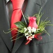 Intimate close-up of a stylish buttonhole fixed to the lapel of a dark charcoal grey suit, evoking the polished yet romantic atmosphere of a Mill Hill wedding day. The central flower is a partially opened rose bud in deep, cerise-pink, its petals velvety and tightly layered, catching the light as they curve inward toward the heart of the bloom. Surrounding this rose, delicate clusters of white baby's breath form a soft, airy frame, their tiny blossoms providing a gentle contrast to the richer pink tones. A ring of green foliage adds depth and natural texture: sturdy, glossy leaves give the arrangement shape, while slender, feathery fronds reach beyond the edges, suggesting the fresh, leafy character of Mill Hill's gardens and parks. At the base, the stems are bound with a bright red satin ribbon tied in a neat bow, its lustrous surface reflecting light and mirroring the strong, clear red of the silk tie beneath the lapel. The suit appears tailored from a fine charcoal wool blend, its subtle texture visible in this close-up, while a grey dress shirt with an understated pattern peeks out from behind, reinforcing the sense of careful coordination. The lighting is soft and even, allowing each surface-the rose petals, the baby's breath, the foliage, the ribbon, and the fabrics-to be appreciated without distraction. Overall, the buttonhole feels refined yet heartfelt, the sort of meticulously crafted detail a Mill Hill florist would prepare for a groom, groomsman, or special guest attending a ceremony at a local church or an elegant NW7 reception venue.