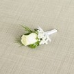 A single white rose boutonniere rests diagonally across a softly textured, linen-like beige fabric, captured in gentle, natural light that feels very much like a calm Mill Hill morning. At the centre, a creamy ivory rosebud is tightly furled, its velvety petals smooth and unblemished, just beginning to open in a way that suggests new beginnings and quiet anticipation before a wedding ceremony. Around the rose, tiny clusters of white baby's breath form airy, cloud-like accents, their minuscule blossoms adding a delicate, lacy halo that softens the whole design. Slender, deep green leaves and a few fine, fern-like sprigs curve around the bloom, providing a rich contrast and a subtle sense of movement, as if gathered fresh from a well-tended garden off Mill Hill Broadway. The stems are neatly bound together with a pristine white satin ribbon, wrapped with visible care and finished in a small, tidy bow that sits just below the flower heads, clearly meant to tuck onto a lapel for a groom or groomsman. The woven, crosshatched texture of the pale fabric background echoes the understated elegance of a classic North London wedding venue, allowing the floral details to stand out without distraction. Soft, even lighting accentuates the gentle shadows between petals, the sheen of the satin ribbon, and the natural gloss of the foliage, creating a refined, timeless feel. The image conveys the intimacy of pinning on a boutonniere before walking into a ceremony, evoking themes of commitment, purity, and the carefully considered touches that make a Mill Hill celebration feel truly personal.