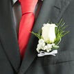 Pinned to the breast pocket of a sharply cut charcoal suit, this white rose boutonniere is shown in close-up, revealing the quiet craftsmanship that might come from a Mill Hill florist preparing flowers on a still morning before guests arrive. At the centre, a single ivory-white rose blooms with soft, layered petals that gently unfurl, their velvety surface catching just enough light to create a halo-like glow. Nestled at its base are dainty sprays of baby's breath, each cluster made up of countless tiny white flowers, forming a delicate cushion that adds volume without overwhelming the rose's calm beauty. Slender fern fronds and looping strands of glossy greenery extend outward, their fresh green tones and graceful curves echoing the natural contours of Mill Hill's tree-lined streets and gardens. The stems are gathered and secured with a neat white ribbon bow, its satin finish smooth beneath the fingers, suggesting both care and tradition. The contrast between the cool grey suit fabric and the warm, living textures of petal and leaf gives the arrangement a sense of balance and refinement. Visually, the boutonniere conveys an air of polished restraint with an undercurrent of emotion, perfect for weddings, proms, or meaningful life events across Mill Hill, from intimate gatherings near the Broadway to more formal venues on the outskirts. Every element works together to turn a simple accessory into a subtle statement of tenderness, optimism, and well-considered style.