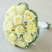 Captured from a slightly raised angle, this classic bridal bouquet gathers approximately fifteen to twenty soft creamy-yellow roses into a plush, rounded dome, each bloom opening into a perfect spiral that suggests quiet refinement. Wisps of white Gypsophila float between the roses like early-morning mist, creating pockets of airiness that contrast the substantial rose heads and soften the visual weight. Several narrow, vibrant green leaves-likely bear grass-arc around the outer edge, their graceful curves lending a contemporary silhouette that cradles the flowers. The stems peek from beneath a tightly wrapped white satin ribbon, its smooth knot and cool finish signalling careful hand-tying. The bouquet rests on a pale taupe linen cloth whose subtle weave complements the floral palette and allows the creamy tones to glow in soft, diffused daylight. Texturally, the piece balances velvet-soft petals, the delicate crispness of baby's breath, and the glossy tension of the grass blades, conjuring an impression of fresh scent and gentle warmth. As a local Mill Hill florist I imagine this arrangement placed on a table near Mill Hill Broadway before a ceremony, or carried by a bride walking toward the park; Flowers Mill Hill can recreate this look for weddings, anniversaries, or intimate ceremonies across the area. The overall mood is one of purity, thoughtful craftsmanship, and timeless romance-an understated statement of love shaped with botanical skill and local care.