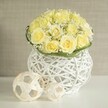 On a pristine white tabletop, a graceful floral centerpiece captures the eye with its dome of light yellow roses, each bloom unfurling in smooth, velvety spirals. The roses are densely packed, forming a rounded silhouette that feels both abundant and carefully controlled, like a cloud of soft sunlight. Threaded among the petals are fine sprays of white baby's breath, their tiny blossoms scattering delicately throughout the arrangement, giving it a light, airy quality reminiscent of blossom catching the breeze over Mill Hill's gardens. Around the base, a slim collar of glossy green foliage encircles the bouquet, adding a fresh, botanical line that neatly separates the mass of flowers from the vessel below. The arrangement is cradled in a distinctive white woven vase, created from slender strands that cross and loop to form an open lattice, similar to contemporary rattan décor often seen in stylish Mill Hill interiors. Light filters through this weave, shaping gentle shadows on the table and enhancing the sense of depth and craftsmanship. To the side, two natural-toned, open-work spheres rest together, their rattan-like strands forming loose, geometric patterns that echo the texture of the vase and complete the modern, organic look. The background is a softly muted beige, slightly textured but out of focus, ensuring the floral design remains the star. The overall impression is one of quiet luxury and refined celebration, ideal for wedding tables in Mill Hill, engagement dinners near Mill Hill East, or as an elegant centerpiece delivered by Flowers Mill Hill to homes and venues across this leafy North London area.