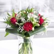 A luxurious red and white bouquet stands proudly in a clear cylindrical glass vase, its stems neatly aligned and visible through the fresh water, like a little slice of Mill Hill sunlight brought indoors. Deep red roses, their velvety petals just beginning to unfurl, cluster near the centre, exuding classic romance and a hint of drama. A large, ruffled crimson carnation anchors the front, its intricate layers adding rich texture and drawing the eye in the way a focal bloom should on a smart dining table off Mill Hill Broadway. Scattered between the reds are pure white and creamy flowers: elegant white alstroemeria with slim, speckled petals, plump white chrysanthemum-style blooms, and several tightly closed white lily buds that promise to open over the coming days. Airy clouds of white gypsophila, or baby's breath, drift throughout the design, softening the strong colours and giving the bouquet a gentle, romantic haze. Around the base, broad, glossy dark green tropical leaves are artfully curved and wrapped, forming a polished collar that frames the flowers. Finer, eucalyptus-like greens and pointed leaves weave in and out, adding depth and fresh woodland tones. The entire arrangement rests on a clean white surface, set against a softly blurred, bright background that suggests daylight filtering through a window, much like the soft light over local gardens near Mill Hill Park. The atmosphere is polished yet warm, a bouquet that feels perfectly suited to anniversaries, special birthdays, Mother's Day, or simply to brighten a Mill Hill home or office with an elegant expression of affection and celebration.