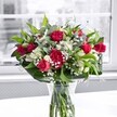 A richly detailed close-up of a romantic red and white bouquet stands in a clear glass vase on a bright white table, with soft daylight filtering through a large, out-of-focus window behind it. At the heart of the arrangement are velvety crimson red roses, some fully unfurled to reveal layers of plush petals, others in elegant bud form promising more beauty in the days ahead. Nestled beside them are full, ruffled red carnations in a matching shade, adding a sense of traditional warmth and extra volume. Threaded between the deep reds are pristine white alstroemeria, or Peruvian lilies, many still in slender bud, while a few partially opened blooms show off their trumpet-shaped petals touched with faint green at the tips. Wisps of delicate white gypsophila, or baby's breath, float between the larger flowers like tiny clouds, giving the bouquet a gentle, airy lightness. Lush greenery forms a graceful frame: broad, glossy deep-green leaves cradle the flowers from below, while slimmer, softer green foliage weaves upward, adding height and movement. The flower stems are clearly visible through the cylindrical glass vase, their cut ends resting in clear water that speaks of freshness and careful preparation. The white tabletop casts a faint reflection at the vase's base, enhancing the clean, modern feel. Behind, the window reveals only a blurred, almost monochrome glimpse of Mill Hill beyond, perhaps recalling a quiet residential street or a park on a muted day, which makes the bouquet's reds, whites, and greens appear even more vivid. The overall mood is joyful and polished, ideal for brightening a home in Mill Hill, celebrating an anniversary, or sending a heartfelt thank-you through local flower delivery.