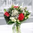 A contemporary mixed bouquet sits in a clear glass vase, captured in soft daylight as if placed beside a bright window in a Mill Hill home. At the heart of the arrangement, a single coral-red rose and a plush crimson carnation provide a rich, velvety focal point, their overlapping petals full and expressive, like a quiet promise or an unspoken thank you. Around them, clusters of delicate white blooms-tiny daisy-like flowers and compact button chrysanthemums-float with a cloud-soft lightness, easing the intensity of the reds and adding a gentle, almost playful rhythm to the design. Creamy white lilies are woven through the bouquet, some just beginning to open, others still in smooth, tapered buds, suggesting new beginnings and beauty yet to come. The stems rise from a simple, cylindrical glass vase, the water clear and cool, catching glimmers of light the way a stream might in Mill Hill Park on a bright morning. At the base, deep emerald leaves have been skillfully folded and wrapped to cradle the flowers, creating wide arcs of glossy green that frame the blooms and give the whole piece a modern, architectural silhouette. The textures range from satin-soft rose petals to feathery daisy centers and the polished surfaces of the foliage, inviting the imagination to reach out and touch. The overall mood is uplifting and elegant-a fresh, thoughtfully balanced display that would feel at home on a dining table overlooking Bittacy Hill, marking a birthday, a graduation, or simply adding a sense of everyday celebration to a quiet evening in NW7.