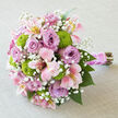 Captured on a softly textured, off-white linen backdrop, this bouquet lies gracefully on its side, revealing its domed, hand-tied structure and rich interplay of colour and texture. The focal point is a cluster of full-bloom lavender roses whose petals spiral outward in velvety layers, each flower forming a generous cup of gentle mauve and lilac hues. Between them, pink alstroemeria stretch out elegantly, their petal tips tinged with blush and ivory, and their centres dusted with subtle yellow and fine strokes of deeper pigmentation that add intricate patterning. Bright green button chrysanthemums punctuate the design like lively exclamation marks, their perfectly round heads composed of tiny, compact petals that provide a fresh, modern edge to the traditionally romantic roses. Threaded all through the arrangement, delicate white gypsophila creates a veil of airy blossoms, adding softness and a sense of movement, as though a light breeze had brushed through a garden in Mill Hill East. The stems of the bouquet are tightly bound, wrapped in a lustrous pink satin ribbon that lies flawlessly smooth, suggesting the practiced hands of an experienced florist at work. A slender twirl of small white pearls is coiled along the handle, catching hints of light and echoing the feel of classic bridal accessories worn at ceremonies near Mill Hill's churches or civil venues. Below the wrapping, the stems fan out in a neat cluster, their natural textures preserved to keep a rustic yet polished appearance. The entire bouquet gives off an impression of delicate fragrance, romance, and celebration, perfectly suited for wedding aisles, engagement parties, or thoughtful deliveries to loved ones across Mill Hill and the surrounding neighbourhoods.