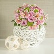 A bright, serene centerpiece evokes the gentle spirit of Mill Hill in this beautifully styled arrangement, perfectly set on a clean white table below a softly textured beige background much like that of quiet Mill Hill Village cafés. The star is a contemporary white vase crafted from slender, woven strips, its openwork design blending modern artistry with homely familiarity found in local handmade décor. The arrangement is lushly abundant with pastel pink and lavender roses-each bloom full and velvety-interspersed with graceful pink alstroemeria, their trumpet shapes delicately streaked for natural variation. Accents of dazzling lime green button chrysanthemums burst joyfully through the arrangement, while fine sprays of white baby's breath dance between the blooms, bringing lightness and movement. Dark green leaves punctuate the ensemble with verdant freshness, tying all the elements together. At the left edge, two decorative spheres fashioned from pale, woven material complete the design, their varying sizes and open patterns reminiscent of objects found at Mill Hill market stalls. The interplay of textures, colors, and organic shapes, all gently illuminated in natural light, creates a centerpiece brimming with warmth and cheerful elegance-ideal for adding a signature flourish to a wedding reception, stylish dinner, or local celebration in and around Mill Hill.