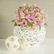 In this close-up scene, a beautifully rounded bouquet takes centre stage, radiating the sort of polished charm that suits a Mill Hill wedding breakfast or engagement celebration near Mill Hill Broadway. The arrangement is carefully composed into a domed silhouette, every stem angled just so to create a seamless blend of colour and texture. Soft lavender and rosy pink roses form the main body of the bouquet, their petals unfurling in overlapping layers that give a plush, almost cushion-like appearance. Nestled between them, pale pink alstroemeria stretch their long, silky petals outward, each bloom displaying the signature speckles and fine streaks of deeper pink and peach that give Peruvian lilies their distinctive character. Adding a vivid counterpoint, lime green button chrysanthemums are tucked throughout the design, their small, perfectly rounded heads lending structure and a bright, modern freshness that lifts the pastels around them. Airy sprigs of tiny white baby's breath thread through the arrangement, softening the transitions between each flower and creating a cloud-like lightness. Dark green foliage peeks out subtly from beneath, framing the bouquet and providing contrast. The flowers are gathered into a unique, spherical vase crafted from white woven wicker or rattan, its open latticework pattern catching the light and introducing a tactile, sculptural quality. This globe-shaped container rests on a pristine white table or shelf that gently reflects the shapes above. Nearby, two smaller decorative spheres made from a paler, natural woven material echo the same open, airy pattern, their broader strips adding a slightly different rhythm to the texture. Behind everything, a neutral wall in soft beige or off-white remains quietly in the background, ensuring all attention rests on the floral artistry. The overall impression is one of gentle sophistication and joyful romance, ideal for a curated wedding flower collection in Mill Hill.