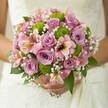 Brimming with gentle charm, this Mill Hill bridal bouquet rests in a bride's hands, its vivid blossoms framed by the subtle textures of an elegant white gown. The arrangement is a tapestry of soft lilac and blush pink roses-some petals still glistening with morning dew-interwoven with crisp green button chrysanthemums for a playful, lively touch. Alstroemeria flowers, painted in pink with maroon flashes and hints of yellow, add intriguing detail, while feathery sprays of baby's breath lend a cloud-like, ethereal softness to the scene. Underneath, deep green foliage offers a grounding contrast to the pastel petals, framing the spherical silhouette of the bouquet with natural grace. Sunlight flowing through a nearby Mill Hill window highlights every nuance of petal and leaf, bringing the bouquet to life and capturing the sense of a new beginning. The overall mood is one of understated luxury and fresh celebration, echoing the spirit of weddings throughout Mill Hill-whether exchanged under the old trees at Mill Hill Park or in the historic halls nearby. Crafted with local expertise, this bouquet is a luminous tribute to timeless romance and community joy, perfect for wedding features or inspiration.