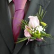 Bathed in gentle natural light, this elegant buttonhole arrangement tells a story of careful craftsmanship and the spirit of celebration unique to Mill Hill. A single pastel pink rose, its petals full and velvety, sits atop the crisp lapel of a deep charcoal suit, standing as a symbol of admiration on a day of joy. Nestled beside, a delicate bloom-likely an alstroemeria-displays streaks of playful magenta, adding spirited flourish reminiscent of walks in Mill Hill Village. Airy clouds of baby's breath encircle the main flowers, suggesting both innocence and timeless romance. The green button mum, bright and tightly clustered, introduces a contemporary pop, brought to life by feathery fronds and slender greenery, evoking the soft texture of local meadows. The creation is thoughtfully secured with a pale lavender ribbon bow, its shimmer echoing the silk of the wearer's striking magenta tie below. The scene evokes both confidence and tenderness-the kind found in milestone moments celebrated across Mill Hill. A testament to the pride and artistry of local florists, this boutonniere elevates any formal occasion, inviting those present to savour the harmony of colour, fragrance, and heartfelt care.