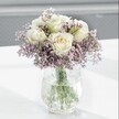 A close-up photograph shows a tranquil, handcrafted bouquet of creamy white roses surrounded by a cloud of soft lavender-purple statice, resting in a clear crackle-glass vase on a pure white tabletop. Each rose is fresh and full, its petals unfolding in gentle spirals, with a faint blush of pale yellow at the centre that hints at early-morning opening. They sit slightly taller than the other blooms, creating a serene focal point that draws the eye to their velvety texture and classic shape. Around them, countless tiny statice flowers form a misty halo of lilac and lavender tones, their papery petals clustered along fine stems that add airiness and delicate texture. The vase itself, textured like frozen water, catches the daylight that filters in from one side, scattering pinpoints of light across its uneven surface and creating subtle reflections on the smooth, white surface below. Through the clear water, straight green stems are visible, crisply cut and neatly arranged, showing the quiet skill of an experienced florist. The softly blurred background melts into gentle shades of grey and off-white, ensuring the bouquet remains the undisputed star of the image, calm and composed. The overall mood is peaceful and reflective, evoking the feeling of a quiet corner in a Mill Hill home, perhaps near a window overlooking local greenery. It is the kind of arrangement you might send to offer comfort, to celebrate a thoughtful birthday, or to bring a sense of understated elegance into a study or bedroom, where its soothing colours and simple beauty can be quietly appreciated throughout the day.