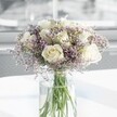 A generously full, round bouquet of creamy white roses and soft lavender statice stands in the centre of the image, arranged in a tall clear glass cylinder vase on a bright white surface. Each rose is a perfect bloom, petals unfolding in layers of ivory and warm cream, with just the faintest hint of blush at the heart, creating a feeling of calm, classic elegance. Around and between the roses, clouds of tiny lilac-purple flowers, likely statice, form a delicate haze, their papery, fern-like sprigs adding fine texture and a gentle mist of colour that softens the arrangement. The green stems are visible through the glass, cut cleanly and standing in crystal-clear water, giving a glimpse of the florist's careful craftsmanship. Soft, natural daylight pours in from a blurred window background, typical of a serene Mill Hill sitting room or conservatory, making the petals appear almost luminous and casting the faintest shadows on the smooth table below. The overall mood is tranquil and uplifting, as if the bouquet has just been delivered fresh from a local Mill Hill florist for a quiet anniversary breakfast, a comforting sympathy gesture, or to bring simple luxury to a modern home. The composition feels airy yet structured, with no additional greenery to distract from the interplay of white and lavender, emphasizing purity, tenderness, and quiet affection. It is the sort of refined rose arrangement that would sit beautifully in a hallway overlooking Mill Hill Park, radiating grace without ever seeming fussy or overstated, and inviting you to imagine the soft, nostalgic scent of roses mingling with the freshness of just-cut stems.