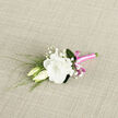 Set against a softly textured, neutral fabric, this boutonniere appears as if it has just been finished on a florist's bench in Mill Hill, ready to be collected for a special day. At its centre sits a lush white lisianthus bloom, fully open and filled with velvety, ruffled petals that spiral outward, creating an impression of calm elegance and quiet romance. The shade of white leans toward creamy ivory, lending warmth rather than starkness, perfect for complementing classic suits or more modern attire alike. Nestled to one side are two lisianthus buds, still gently closed and resting close together, suggesting companionship, unity, and the start of a shared journey. Threaded between the main flower and buds are wisps of baby's breath, their diminutive white blossoms clustered along fine stems, adding a light, airy texture, almost like a dusting of soft snow. Slender, grass-like leaves and thin green stems arc gracefully around the arrangement, providing contrast and a fresh-from-the-garden feel that recalls strolls through nearby green spaces like Mill Hill Park or Arrandene Open Space. The stems are meticulously gathered and encased in a bright pink satin ribbon whose smooth, glossy surface shimmers slightly in the light. A neat, compact bow completes the wrap, giving the design a touch of playfulness while keeping it refined. The weave of the background fabric adds subtle visual interest without competing with the flowers themselves. Overall, the image conveys a sense of intimacy and care, showcasing a boutonniere ready for a wedding, prom, or special celebration anywhere in and around Mill Hill.
