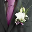 Pinned close to the chest on a dark, finely tailored jacket, this wedding boutonnière feels like a refined nod to tradition, ideal for a groom celebrating in Mill Hill. The focal point is a single white lisianthus bloom, fully opened and beautifully formed, its layered petals soft and velvety to the eye, curling delicately outward like carefully pressed crepe. Behind the main flower, two lisianthus buds rise on slim stems, their pale green sheaths still protecting the petals within, suggesting the quiet promise and potential that accompany a wedding day near Mill Hill East or along the quiet residential streets by Lawrence Street. Surrounding the central bloom, airy clusters of baby's breath add volume without heaviness, each tiny white flower forming part of a mist-like ring that imparts a touch of romance and lightness. Slender blades and sprigs of green foliage thread through the design, offering slight shifts in tone and texture that recall the varied planting in local gardens and park borders. At the base, a single deep-green leaf is gently curled or folded, its glossy surface forming a polished cradle that frames the white lisianthus and keeps the boutonnière visually grounded. The stems are neatly wrapped in a narrow mauve ribbon, finished with a small, precise bow whose lavender hue subtly complements many wedding colour schemes popular in North London. The overall effect is one of poised elegance and calm celebration, as though the piece carries a faint, fresh floral scent that lingers delicately in the air with every movement of the groom throughout the ceremony and reception.