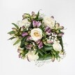 An overhead view of a hand-tied bouquet shows creamy white and soft ivory roses at different stages of bloom, resting against a perfectly clean white background that makes every petal stand out with clarity. Some roses are still gently cupped around a pale buttery centre, while others are fully open, their velvety petals spiralling outwards in generous layers that suggest quiet romance and care. Threaded between the roses are graceful stems of alstroemeria, their slender buds and half-open flowers revealing a blend of mauve, lavender, and purplish-pink tones, brushed with fine green streaks that give each bloom a painterly character. Wisps of gypsophila, or baby's breath, cluster like tiny white clouds, softening the spaces between the larger flowers and adding an airy, almost weightless texture. A collar of rich, deep green foliage frames the bouquet, the broad, glossy leaves folding and curving to hold the blooms as if in an embrace, giving the entire design a sense of freshness and structure. The lighting feels like soft daytime brightness, the kind you might notice through a Mill Hill bay window overlooking Mill Hill Park, highlighting the natural sheen of the leaves and the delicate shadows in each rose. This compact yet full arrangement feels ideal for a thoughtful delivery to a Mill Hill home or office-something to mark a gentle celebration, offer comfort after difficult news, or simply bring refined elegance to a kitchen table. Every detail suggests the experienced hand of a local florist who understands balance, texture, and the quiet joy fresh flowers can bring to everyday life in NW7.