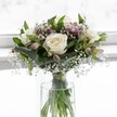 An elegant white rose bouquet stands in a clear cylindrical glass vase, bathed in soft daylight that feels like it could be streaming through a bright Mill Hill kitchen window. Three creamy white roses, fully open and velvety, form the calm heart of the arrangement; their petals spiral outward in gentle curves, with ivory centers that suggest absolute freshness and quiet purity. Around them, polished deep-green foliage is carefully structured, some leaves folded and cupped as if cradling the blooms, giving the bouquet a rounded, composed silhouette. Threads of delicate white gypsophila, or baby's breath, float through the design like tiny clouds, their tiny star-shaped blossoms creating a lace-like veil between the larger flowers. Slim stems of alstroemeria, some still in bud and others just beginning to open, add subtle touches of muted mauve and blush-pink, their hints of colour bringing depth and a whisper of romance without disturbing the predominantly white palette. The stems are visible through the clear water in the vase and appear neatly gathered and tied with a fine ribbon just above the waterline, showing the florist's care and precision. The background is softly blurred and luminous, with no harsh shadows, so the bouquet appears almost to glow, as if poised on a windowsill in a quiet home off Mill Hill Broadway. The overall mood is tranquil and refined, perfect for a thoughtful get-well gesture, a peaceful sympathy tribute, or a simple expression of appreciation delivered locally around Mill Hill, evoking serenity, tenderness, and timeless grace in one compact, beautifully balanced design.