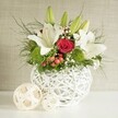 A serene yet striking wedding table arrangement is pictured against a softly textured, light-toned wall, evoking the peaceful character of Mill Hill's tree-lined avenues. At the centre stands a distinctive white spherical vase, constructed from overlapping bands that create an open, woven pattern reminiscent of delicate basketry rendered in smooth ceramic. From this sculptural vessel, a lush mix of flowers and foliage spills outward with graceful ease. Large white lilies dominate the design, their petals spreading wide in elegant star shapes, each bloom revealing delicate interior speckles and golden stamens that catch the light. Around them, several green lily buds rise upright, suggesting that the arrangement will continue to open and evolve over the course of a wedding reception or special dinner. Nestled close to the lilies are velvety red roses, with one particularly prominent flower in the centre, its tightly layered petals curving inward to form a classic romantic spiral-perfect for symbolising devotion at a Mill Hill wedding or anniversary celebration. Between the lilies and roses, bright green pompon blooms add rounded bursts of colour and a soft, tufted texture that echoes the circular form of the vase. Fine sprays of white baby's breath weave through the bouquet, creating a misty, cloud-like effect, while small clusters of red berries supply subtle festive notes, ideal for winter gatherings. Wispy fern-like greens and darker leafy stems cascade gently over the rim, adding movement and volume, as if arranged by a florist who understands Mill Hill's love of natural, garden-inspired styling. To the left, two smaller decorative spheres in pale woven material rest on the tabletop, mirroring the vase's open-weave design and completing the composition. The overall impression is of an elegant, contemporary centrepiece perfectly suited to wedding tables and intimate celebrations throughout Mill Hill.
