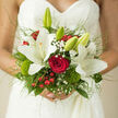This bridal bouquet is shown nestled against the fine lace of a wedding dress, its colours and textures radiating softly, as if filtered through the calm atmosphere of a Mill Hill afternoon. Large white lilies take centre stage, their broad, star-like petals unfolding in smooth, creamy layers that seem to glow in the gentle light. Among them, a single deep red rose offers a bold contrast, its velvety petals spiralling inward, rich and saturated, symbolising enduring love. Threaded throughout are delicate sprays of baby's breath, tiny white flowers that create a misty halo around the more prominent blooms. Bright red berries and small, mossy green clusters bring unexpected texture and cheerful contrast, reminiscent of berries and foliage seen along the paths near Mill Hill Broadway. Fine grasses and airy greenery extend outward, creating soft lines that move the eye around the bouquet and give it a natural, slightly untamed edge, rather than a rigid, traditional shape. The stems are tightly and neatly gathered, clearly hand-tied, suggesting the steady hands of an experienced florist who understands how to balance structure with flow. The lace behind the flowers adds another layer of intricate detail, its delicate patterns echoing the complexity of the bouquet itself. Light and shadow play gently across the petals and leaves, highlighting every curve and fold. The overall impression is romantic, elegant, and full of quiet anticipation, a bouquet ready to accompany a Mill Hill bride from the serenity of her preparations to the joyful moment she steps into view of her guests.