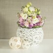 Flooded with soft daylight, this delicate bouquet brings together the joy of spring as celebrated in Mill Hill's gardens and homes. Ruffled lisianthus in creamy white and gentle yellow make up the heart of the arrangement, their blossoms cascading softly from an airy, spherical vase expertly crafted from intertwined white rattan-a nod to North London's love for contemporary craftsmanship. Rising through the bouquet are blush pink stock flowers, whose vertical accents add a lively dance of colour and texture. Anchoring the floral display is a base of vibrant purple fill-perhaps aster or decorative allium-providing a plush contrast to the light blooms above. The entire composition is enveloped in fresh green leaves, their crisp edges mirroring the awakening found in local woodlands each spring. To the left of the vase rest two meticulously woven natural rattan spheres, their open, basket-like intricacy resonating with the vases' hand-finished charm and Mill Hill's tradition of stylish home accessories. The bouquet sits atop a pristine white tabletop, against a tranquil beige wall that softly diffuses the light. This evocative scene from Flowers Mill Hill is ideal for wedding centrepieces, anniversary dinners, or elegant home gatherings across NW7-each element thoughtfully arranged for beauty, peace, and heartfelt celebration.