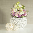 Rounded and refined, this floral design balances softness and structure with the care of a local florist who knows Mill Hill's calmer corners. At its core, creamy white and pale yellow Eustoma (Lisianthus) rise like small lanterns, their layered petals catching soft light; around them, tender pink Stock blooms unfurl in clustered sprays that add a romantic, old-garden scent, while angular purple Allium heads introduce a lively, architectural contrast. The bouquet's foliage carries fresh green notes and is secured at the base with a darker green leaf band that both frames and flattens the silhouette for a composed, abundant look. The vessel is a distinctive spherical white lattice crafted from slender woven branches-an airy, sculptural form that reveals stems and a hint of water and that pairs beautifully with two natural woven orbs placed to the left on the white surface. These wooden spheres and the vase share an organic texture that anchors the pastel palette against a softly textured beige-greige background, reminiscent of a Mill Hill Village sitting room with muted wallpaper or a graceful boutique interior off The Ridgeway. The overall effect feels like a gentle spring sunrise: visually refreshing, quietly fragrant, and comforting. This composition would suit a bedside, a calm office, or a thoughtful gift for someone near Mill Hill Park; it reads as both contemporary design and hands-on floristry, offering tactile rhythm and soothing colour harmony.
