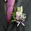 A detailed close-up shows the upper body of a well-dressed gentleman in a dark charcoal suit, the kind you might see outside a Mill Hill registry office or stepping from a car near Mill Hill Broadway on a wedding morning. The suit jacket is tailored and smooth, with a subtle woven texture visible along the lapel and sleeve, and beneath it a crisp, pale dress shirt with a fine grid pattern peeks through. At the centre of the frame sits a vivid magenta silk tie, perfectly knotted, its lustrous surface catching soft, natural light as though the photograph was taken by a window. Pinned to the left lapel, the boutonnière becomes the true focal point: a carefully composed spray of creamy, unopened buds-resembling snapdragons-rising gracefully above clusters of soft pink and lilac blooms that evoke sweet peas or delicate stock. Tiny white tufts of gypsophila add a mist-like softness, while slightly deeper purple florets provide depth and a sense of quiet drama. Around the base, broad, glossy green leaves cradle the flowers, balanced by fine, feathery fronds of brighter foliage that arc gently outward, suggesting movement with every step the wearer takes along the tree-lined streets near Mill Hill Park. A slim lavender satin ribbon is tied neatly around the stems in a precise little bow, harmonising with the lilac tones in the flowers and adding a refined finishing touch. The overall effect is one of romantic, modern elegance-a buttonhole that feels both personal and celebratory, perfect for a groom or honoured guest arriving at a North London wedding, carrying with it the promise of joyful moments ahead.