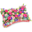 A lovingly made rectangular floral 'pillow' is photographed from a slightly elevated, oblique angle against a crisp white background, the composition emphasizing its soft, cushion-like profile and dense surface of blooms. The top is a mosaic of hot pink roses-some tightly furled, others just beginning to unfurl-whose velvety petals read like satin under the soft studio light. Interwoven are clusters of pale blush spray carnations, their ruffled edges giving the surface a plush, tactile quality. Bright lemon-yellow ranunculus punctuate the design with sunny, buttercup-like warmth while deep purple lisianthus (eustoma) add bell-shaped depth and cool contrast. Silver-blue eryngium thistles introduce a dramatic, spiky architectural note that offsets the softer forms, and small pink filler flowers scatter like gentle confetti across the composition. Rounded, silvery eucalyptus leaves and varied glossy greenery form a verdant foundation beneath and between the flowers, adding scent and a calm, cooling tone. Encircling the base is a rich, deep red pleated satin ribbon, ruffled into a defined border that frames the cushion and lends a subtle formality suited to remembrance. The overall palette-fuchsia, violet, lemon and blush-feels celebratory rather than somber, suggesting warmth, cherished memories and quiet comfort. As a sympathy or memorial tribute this pillow balances artistry and tenderness: you can almost sense the faint, sweet fragrance of the roses and eucalyptus, the soft give of petals beneath fingers, and the careful hand that has arranged each stem. The effect is both consoling and refreshingly bright, a floral embrace intended to honour and remember a life with colour and care.