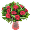 A classic, museum-clean photograph of a lush bouquet built around a dozen or more deep crimson roses, each bloom fully open to reveal tightly spiralled, velvety petals that read almost wine-dark at the edges. The rose heads form a rounded, opulent dome, their rich red surfaces catching soft, even light that reveals subtle veining and the satiny sheen of fresh petals. Nestled between and beneath the roses is an abundance of dark green foliage: glossy flat leaves that create a dense verdant backdrop and larger, brighter green blades artfully folded and rolled into tubular spirals. Those sculpted leaves - evocative of Aspidistra and Salal used by experienced florists - loop and curl like living ribbons, adding modern architecture to a timeless red-rose silhouette. The arrangement sits in a tall, translucent ruby-red glass vase with a faint ribbed or twisted texture; the vase's vertical lines pick up reflections and echo the vertical stems, giving the whole composition a slender, elegant profile. A crisp white background isolates the bouquet, intensifying the contrast between the saturated reds and deep greens and making the composition read as both intimate and celebratory. There is an implied scent of classic rose perfume in this image, a comforting, romantic warmth that suits anniversaries, Valentine's gestures, or a refined centrepiece for a special dinner. As a local Mill Hill florist might suggest, this is the sort of arrangement that sits perfectly in a bright flat on Mill Hill Broadway or as a striking welcome on a mantel in Mill Hill Village, where the colours read as both modern and timeless.