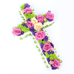 Against a spotless white backdrop, this floral cross stands out as a vibrant yet reverent sympathy tribute, angled slightly from upper left to lower right so its texture and form can be clearly seen. The shape of the cross is carefully outlined with a dense border of white spray chrysanthemums, each small bloom contributing to a soft, cloud-like perimeter. Just beneath this white frame, a slim strip of pale green ribbon or meticulously folded foliage surfaces around the edges, providing a touch of refined colour and echoing the fresh greenery of Mill Hill's gardens and open fields. Within this border, the centre of the cross is lavishly filled with an array of flowers that create a harmonious but striking palette. Large roses in intense fuchsia pink are scattered throughout, their fully opened petals forming generous focal points, while companion roses in creamy pale yellow or peach lend warmth and tenderness. Interspersed among them are rounded lime-green button chrysanthemums, their tight, almost pom-pom-like heads adding cheerful texture and a contemporary note. Woven between the larger flowers are clusters of smaller deep purple and violet blossoms-likely statice or lisianthus-that introduce depth and a more contemplative tone. The overall design appears full but not overcrowded, allowing each flower type to be appreciated. Bright, even lighting casts no harsh shadows, so every petal, ribbon fold, and colour transition remains clearly visible. This thoughtfully crafted floral cross, ideal for a Christian funeral, memorial, or graveside tribute in Mill Hill, conveys both the solemnity of loss and the enduring comfort of beauty and faith expressed through fresh, carefully chosen blooms.