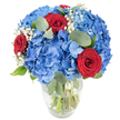 A lush, tightly rounded bouquet sits in a clear glass vase with a slightly rounded base and a gently tapered neck, the stems visible through sparkling water. Dominating the arrangement are large, cloud-like clusters of sky-blue hydrangeas whose overlapping petals form a soft, voluminous mass that feels cool to the eye. Nestled among the blue are three deep red roses, their velvety petals still tightly furled so each bloom reads as a rich, concentrated point of passion against the softer azure. Scattered throughout are tiny, ivory sprays of baby's breath that lift the composition with an airy, starry texture, like breathy little clouds caught between the larger blooms. Muted green eucalyptus leaves - round, silvery and matte - weave through the flowers, adding organic depth and a faint herbal note you can almost smell. The bouquet is lit in bright, natural light against a clean white backdrop, which sharpens the saturated blue and red while underscoring the freshness visible in the submerged stems. As a florist in Mill Hill I imagine this piece on a long wooden table near The Ridgeway, bringing a celebratory, quietly patriotic mood to anniversaries, birthdays, or a dinner among friends. The balance of plush hydrangea mass, pointed rose drama, and delicate gypsophila detail creates a composition that soothes and enlivens at once - a confident, contemporary arrangement that reads as both romantic and joyful without being fussy.