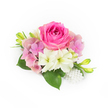 A compact, bridal-ready corsage rests against a pristine white background, every element arranged with careful restraint and quiet elegance. At its centre sits a lush bubblegum-fuchsia rose, its tightly swirled inner petals opening outward in velvet layers that catch the soft, natural light; you can almost imagine the rose's subtle, fresh fragrance. Encircling the rose are cushiony clusters of pale pink and lavender hydrangea florets, their papery textures creating a feathery halo that softens the composition and lends a romantic, vintage air. Two creamy white, trumpet-shaped blooms-reminiscent of tiny amaryllis or lilies-tilt gently below the main flower, revealing pale green throats that add a cool, luminous contrast. Deep, glossy green leaves fan out beneath the blooms, their broad surfaces grounding the piece and giving it a garden-fresh weight. Slim, vibrant green buds with delicate yellow tips punctuate the sides, suggesting new life and upward movement as if the corsage were still growing. A small cluster of pearlescent beads cascades at the lower right, varying subtly in size and catching light like heirloom jewellery sewn into the design; this refined touch makes the piece feel especially suited to proms, wedding corsages, or intimate anniversary moments. The overall texture moves from velvet to airy to satin, and the arrangement sits in a halo of clean daylight ideal for photographs or close viewing. Crafted with the sensibility of a local florist who knows Mill Hill tastes, this compact floral detail is a statement of gentle celebration and timeless femininity, perfect for spring and summer occasions around Mill Hill Village and nearby green spaces.