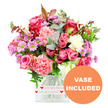 This photograph captures a lush, optimistic bouquet brimming with colour, displayed in a simple yet elegant clear glass vase with a square, softly rounded shape, all set against a clean white background. The flowers form a full, rounded arrangement dominated by shades of pink: deep fuchsia roses with richly coloured petal edges sit alongside softer blush-pink roses, each bloom layered and velvety. Between them, generous carnations in coral, peach-pink, and creamy white lend their ruffled texture, giving the design a plush, comforting feel, as if it has been arranged to brighten a living room in Mill Hill on a grey day. Smaller daisy-like chrysanthemums peek out here and there in lavender and gentle purple hues, with luminous lime-green centres that add a cheerful spark and ensure the arrangement never feels flat. The bouquet is surrounded by a collar of mixed greenery-cool-toned eucalyptus leaves, deeper green sprigs, and a light scattering of reddish-brown foliage that introduces a hint of romantic warmth, echoing the colours you might notice on an evening walk around Mill Hill Park in late summer. The clear glass vase is finished with a decorative white band wrapped around its base, adorned with repeating red hearts and the sentimental phrase all my love, clearly suggesting this is a bouquet meant to send affection, whether for a birthday, an anniversary, or a simple thinking of you to someone near Mill Hill Broadway. In the lower right-hand corner of the image, a bold orange circle bearing the words VASE INCLUDED in crisp white capitals stands out, highlighting that the arrangement arrives as a complete, ready-to-enjoy floral gift for homes and offices throughout the Mill Hill area.