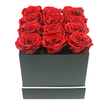 The image presents a stylish floral gift box that embodies modern romance, the sort of design a Mill Hill florist might recommend for a meaningful, elegant gesture. At the centre sits a squared, matte black box with clean, architectural lines and a smooth, understated finish that absorbs light. Around its lower edge runs a narrow horizontal band of white, a subtle decorative touch that brings a touch of contemporary flair, echoing the minimalist interiors of many homes off Hale Lane and around Mill Hill Circus. Emerging from the top opening are twelve sumptuous red roses, meticulously arranged in three uniform rows of four. The blooms are pressed closely together, creating a continuous surface of crimson that gently swells above the rim of the box. Each rose is in full bloom, its velvety petals unfolding in tight spirals that form intricate, layered rosettes. The colour is a deep, luxurious red with delicate shifts in tone, from darker shadows at the heart of each flower to slightly brighter highlights at the edges of the petals. The roses appear unusually consistent in size and perfection, suggesting they may be preserved, chosen for their ability to stay beautiful long after they arrive. The entire arrangement is photographed against a pure white background, with no other objects in view, so the focus remains firmly on the striking contrast between the dark box and the vivid roses. Soft, diffused lighting caresses the scene, revealing gentle shadows along the petal folds and the box's base, adding dimension without harshness. The effect is both intimate and grand, ideal for a romantic delivery to a home near Mill Hill Park or a surprise waiting on the table after dinner in a quiet Mill Hill Broadway apartment.