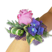 A wearable jewel of floral craftsmanship rests on a fair-skinned wrist against a clean white backdrop, presented like a small keepsake of summer evenings. At its heart sits a partially open pink rose, the petals soft and layered, their edges shaded toward a richer magenta that catches the light like velvet. Beside it, tight clusters of deep indigo to purple hydrangea lend a dramatic counterpoint; each tiny floret shows a faint, cool blue centre that seems to glow against the rose's warm blush. The composition is grounded with slender, silvery-green sprigs-suggestive of lavender or eucalyptus-whose fine texture brings an airy, herbaceous lift and a hint of fresh, mineral fragrance. Variegated English ivy frames the blooms, glossy leaves dark at the centre and rimmed with creamy yellow, introducing a classic, garden-like formality. A modern accent-a broad green leaf banded with white and light green-is artfully coiled into a tight spiral, adding sculptural movement and a contemporary silhouette. Underpinning the arrangement, an open-weave mesh of deep violet peeks through in lacy layers, amplifying the hydrangeas' purple tone and giving the corsage a subtle play of shadow and depth. The overall impression is refined and celebratory: tactile contrasts of silky rose, velvety hydrangea, crisp ivy and feathery greens invite touch, while the restrained colour palette reads like evening wear for a wedding, prom, or graduation. As a florist who often sources seasonal blooms in Mill Hill and the surrounding lanes, I imagine this wristlet worn along The Ridgeway or at a reception held near Mill Hill Park-an elegant, carefully balanced piece that feels both timeless and freshly modern.