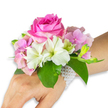 Soft studio light falls across a wrist corsage gently resting on the left hand and forearm, set against a clean white backdrop that makes the colours sing. At the centre sits a large, fully opened deep fuchsia-pink rose - petals densely layered, velvety to the eye and curling outward in a confident spiral. Surrounding the rose are clusters of blush-pink hydrangea florets that add a cloud-like, textured base and a delicate volume; their petals hold a translucent, dewy quality. Interspersed among these are pristine white alstroemeria (Peruvian lilies), each with four to six slender petals brushed with pale green streaking toward the throat, lending a crisp, botanical contrast. On the right-hand side a tight cluster of pale green alstroemeria buds and a few nearly closed pink buds promise gentle unfurling, introducing movement to the composition. Lush, broad emerald leaves cradle the blooms, their glossy surfaces and deep veins providing a fresh, grounding backdrop. The arrangement is secured to the wrist with an elegant multi-strand bracelet of small, lustrous white pearls that wrap comfortably and trail slightly across the back of the hand like heirloom lace. You can almost imagine the faint scent of rose warmed by skin, the tender papery notes of lilies, and the cool, leafy green beneath. This refined corsage reads as both classic and youthful - perfect for a prom, wedding, or intimate celebration in Mill Hill, whether being carried down The Ridgeway or enjoyed during a garden photo in Mill Hill Park. Its careful construction shows considered floristry and wearable finesse suited to special moments close to home.