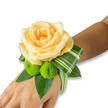 An intimate wrist corsage rests gently on the inner forearm, the centerpiece a buttery yellow rose that opens in luminous, layered petals like the first light of day. Beneath it, two lively lime-green button chrysanthemums - small, spherical pom-poms with tightly packed florets - nestle close, their bright texture offering a cheerful counterpoint to the rose's velvety smoothness. Variegated foliage with crisp white striping is artfully curled to one side, its ribbon-like twist drawing the eye along the wrist and giving the design an architectural grace. Rich, glossy leaves form a deep green base, their broad surfaces reflecting soft studio light and grounding the arrangement with lush depth. The flowers are secured as a delicate wristlet that appears lightweight and wearable, cradling the skin in a way that suggests both comfort and occasion-ready polish. The overall palette - warm buttery yellow, vivid lime green, and layered greens - reads as quietly celebratory, scented with a faint whisper of rose and cut foliage. Worn in anticipation of a wedding vow or evening reception, this wrist corsage communicates gentle excitement and thoughtful craftsmanship, like a small keepsake to be carried through a day of close embraces and photographs. As a Mill Hill floral design, the piece reflects local sensibilities: elegant enough for a ceremony at St. Paul's Church yet relaxed enough for a village dance near Mill Hill Broadway. Freshly made and finished with careful folding of the variegated leaf, it's the kind of floral accessory that holds up under soft light and busy hands, perfect for marking a treasured moment with natural beauty and tactile refinement.