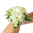 A graceful white wrist corsage rests against fair skin, angled up from the lower right into a clean white frame, its composition quietly refined. At the centre sits a creamy ivory rose, petals unfolding with a velvety softness that catches the light with a warm, muted glow. Beneath and just left of the rose, a larger trumpet-shaped white bloom-resembling an alstroemeria or small lily-reveals a pale green-yellow throat that gives the arrangement gentle depth. Sprigs of tiny, cloud-like white filler flowers, like baby's breath, drift around the main blooms, adding an airy texture that softens the silhouette. Small bright-green buds, some tightly closed, hint at future opening and lend a fresh, hopeful rhythm to the piece, while a cluster of daisy-style white florets with golden centres adds a sweet, cheerful punctuation near the wrist. Broad, glossy deep-green leaves frame and cradle the flowers, their waxy surfaces creating a lush contrast against the pristine whites. The entire arrangement is secured by an elegant multi-strand pearl bracelet; rows of ivory pearls encircle the wrist, their cool smoothness complementing the blossoms and suggesting quiet luxury. The light is soft and diffuse, evoking an early spring morning and a clean, celebratory mood-perfect for weddings, proms, or formal dances. As a Mill Hill floral design, it carries the subtle, handcrafted sensibility of local makers who know how to balance tradition and contemporary wearability, an accessory that reads as both classic and intimately personal for special moments close to home.